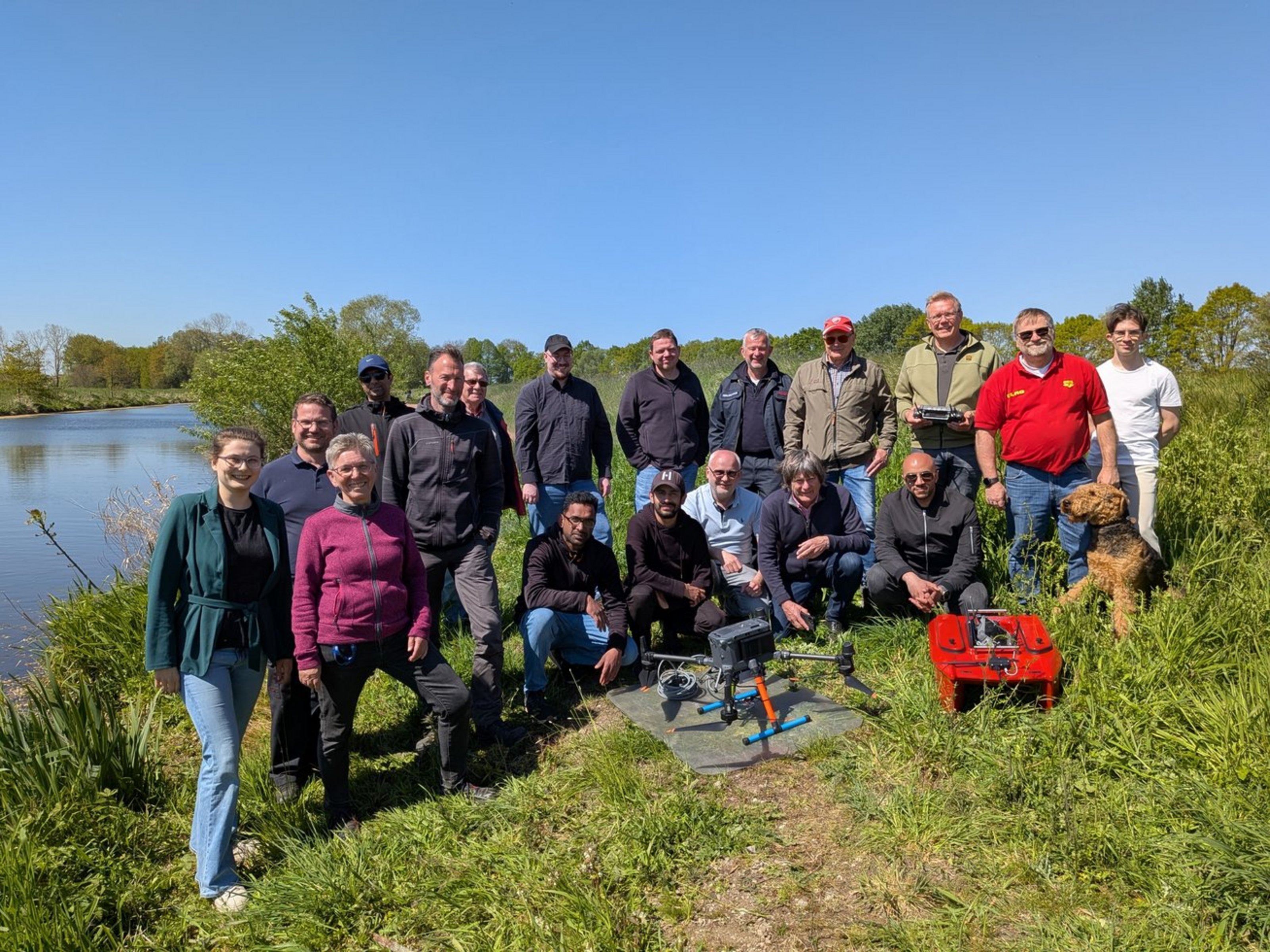 Gruppe von Menschen steht und kniet auf einem grasbewachsenen Weg neben einem See mit Drohnen und einem Hund