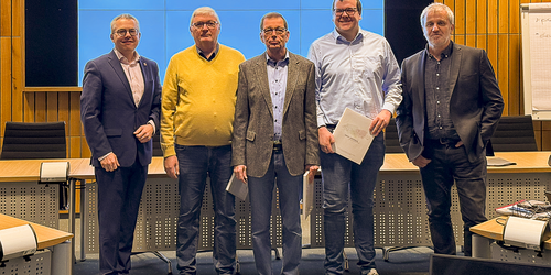 Fünf Männer stehen nebeneinander vor einer Leinwand. Die drei Männer in der Mitte halten eine weißes Buch in der Hand.