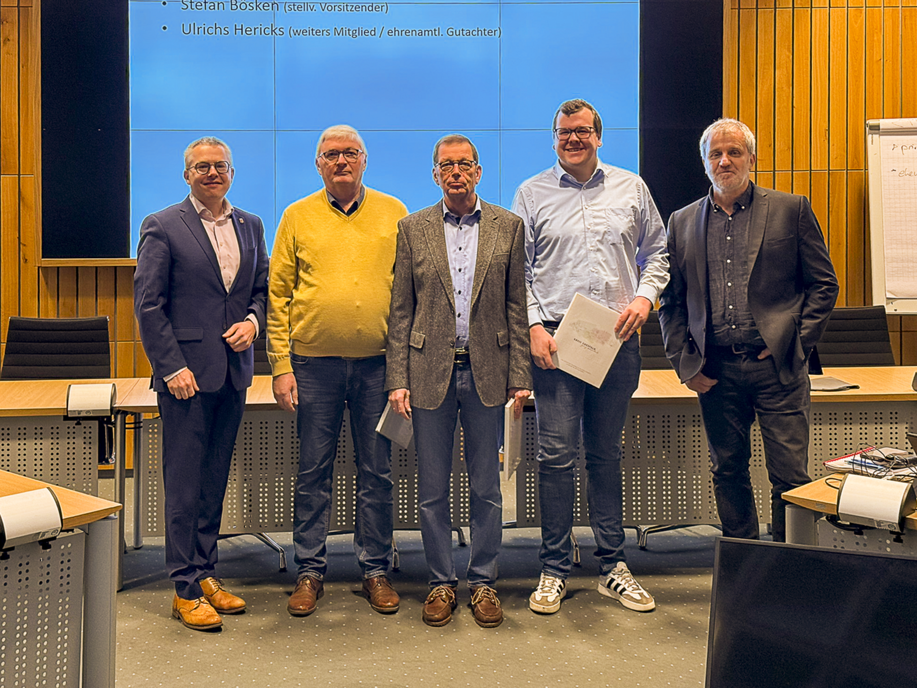 Fünf Männer stehen nebeneinander vor einer Leinwand. Die drei Männer in der Mitte halten eine weißes Buch in der Hand.