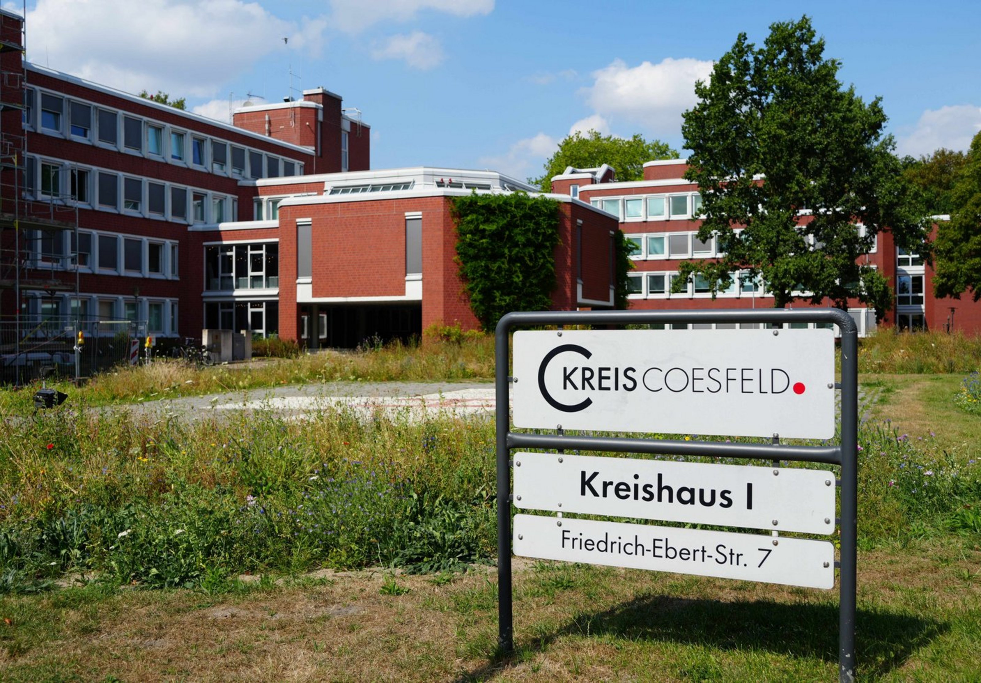 Man sieht ein Schild auf einer Wiese mit der Aufschrift Kreishaus eins. Im Hintergrund ist ein rotes Backsteingebäude.