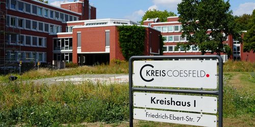 Man sieht ein Schild auf einer Wiese mit der Aufschrift Kreishaus eins. Im Hintergrund ist ein rotes Backsteingebäude.
