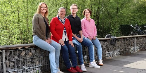 Drei Frauen und ein Mann sitzen auf einer Bank. Hinter ihnen wachsen Bäume.