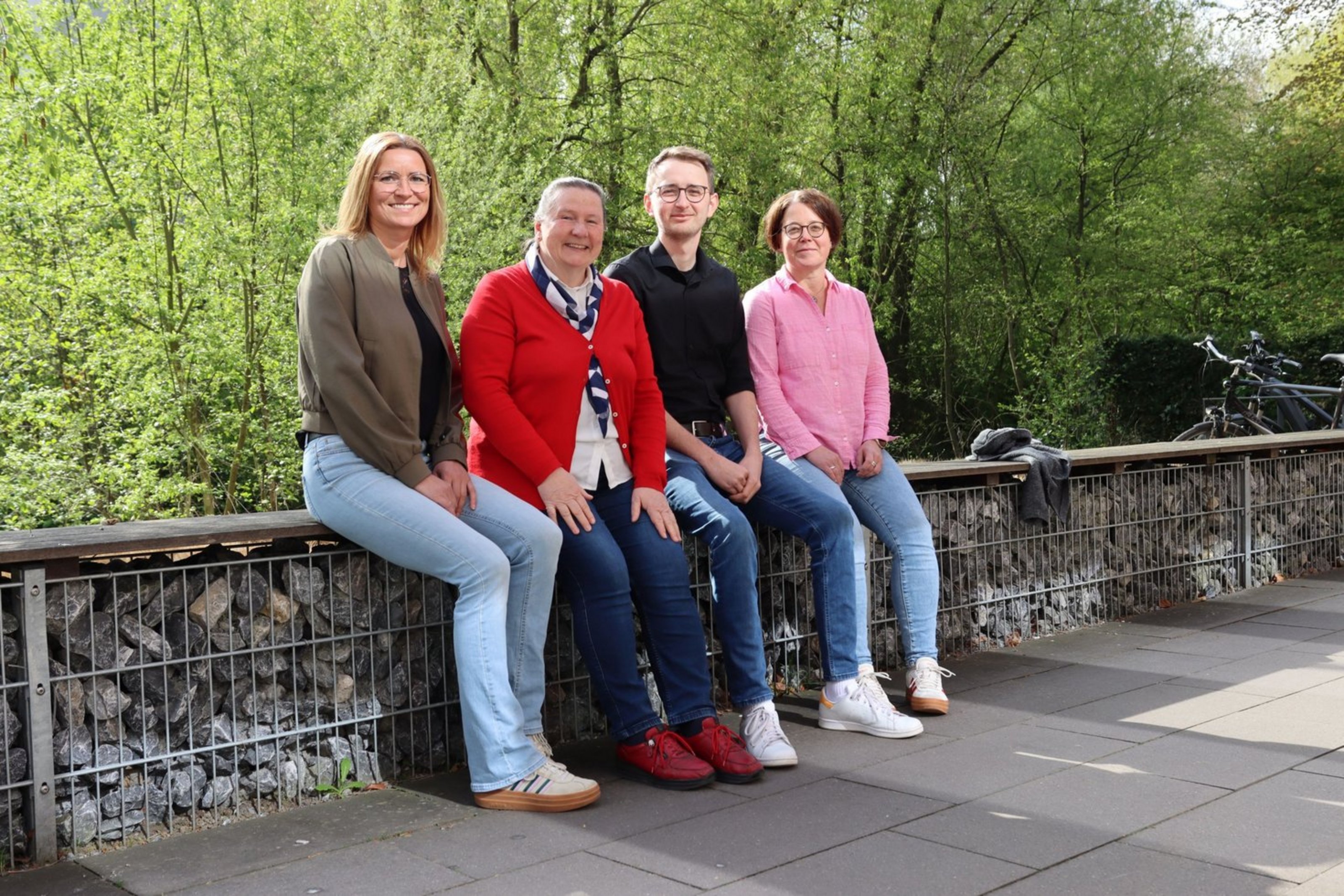 Drei Frauen und ein Mann sitzen auf einer Bank. Hinter ihnen wachsen Bäume.