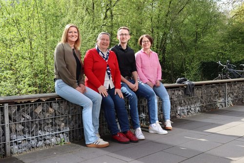 Drei Frauen und ein Mann sitzen auf einer Bank. Hinter ihnen wachsen Bäume.