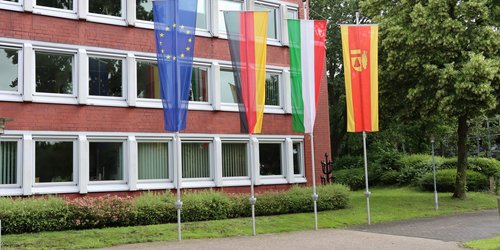 Vor einem roten Gebäude mit vielen Fenstern sind von links nach rechts die Europaflagge, die Flagge der Bundesrepublik Deutschlands, Nordrhein-Westfalens und des Kreises Coesfeld gehisst.