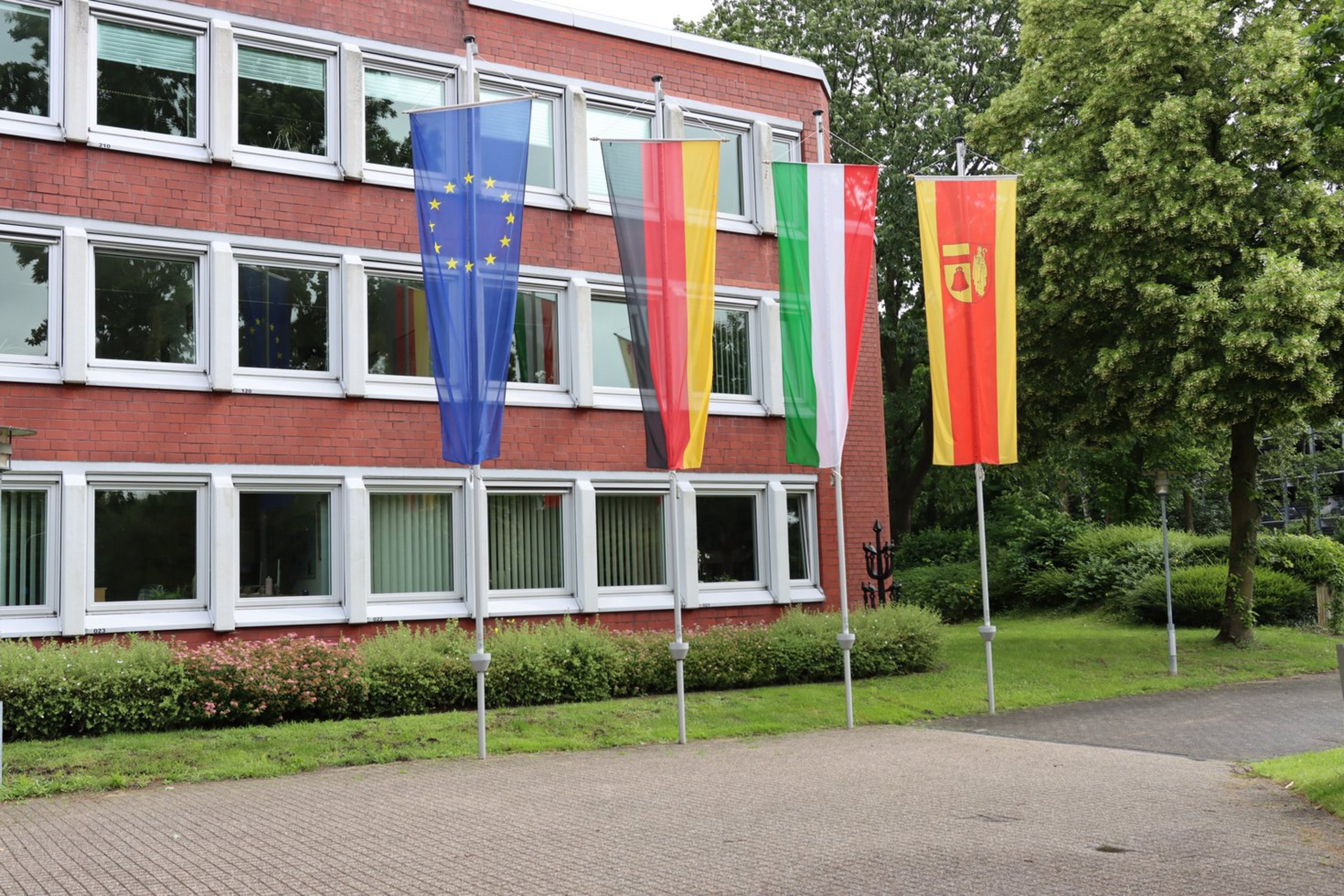 Vor einem roten Gebäude mit vielen Fenstern sind von links nach rechts die Europaflagge, die Flagge der Bundesrepublik Deutschlands, Nordrhein-Westfalens und des Kreises Coesfeld gehisst.