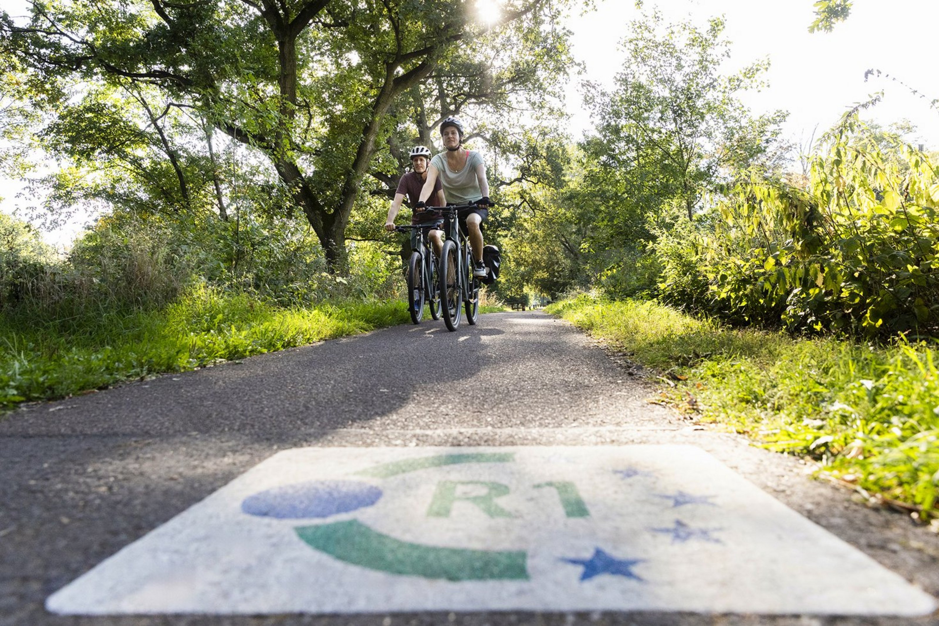 Auf einem Radweg fahren zwei Fahrradfahrer auf die Kamera zu. Der Weg ist umringt von grünen Bäumen und Gebüsch. Auf dem Asphalt ist ein Logo zu sehen mit der Aufschrift R1., 