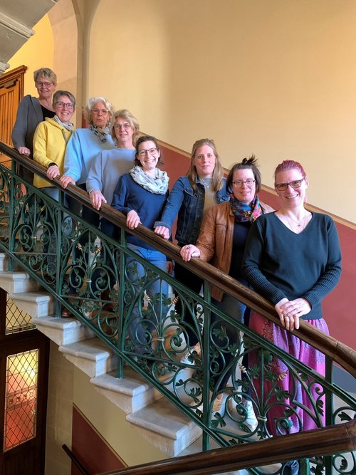 „Acht Frauen stehen auf einer alten, kunstvoll verzierten Treppe mit grünem Geländer in einem historischen Gebäude. Sie blicken freundlich in die Kamera und stehen hintereinander auf den Stufen. Die Gruppe wirkt fröhlich und herzlich, einige tragen bunte Schals oder Brillen. Die Wand im Hintergrund ist in warmen Farben gestrichen und unterstreicht die einladende Atmosphäre.“