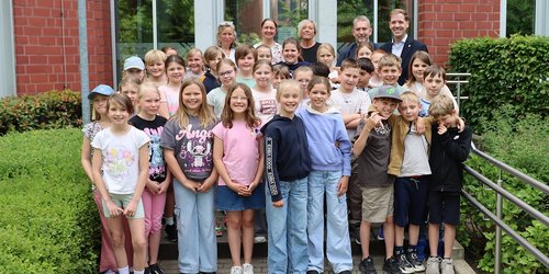 Eine Gruppe von rund 20 Kindern steht vor dem Eingang eines Gebäudes aus rotem Stein. In der hinteren Reihe stehen drei Frauen und zwei Männer