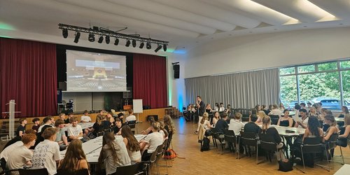 Großer, heller Veranstaltungsraum mit mehreren runden Tischen, an denen etwa 60 Jugendliche in Gruppen sitzen und miteinander arbeiten oder diskutieren. Im Hintergrund befindet sich eine Bühne mit rotem Vorhang, einer großen Projektionsleinwand und Veranstaltungstechnik. Eine moderierende Person steht zwischen den Tischgruppen und spricht mit den Teilnehmenden. Rechts sorgen bodentiefe Fenster für viel Tageslicht und geben den Blick auf Bäume im Freien frei. Die Atmosphäre wirkt offen, konzentriert und gemeinschaftlich.