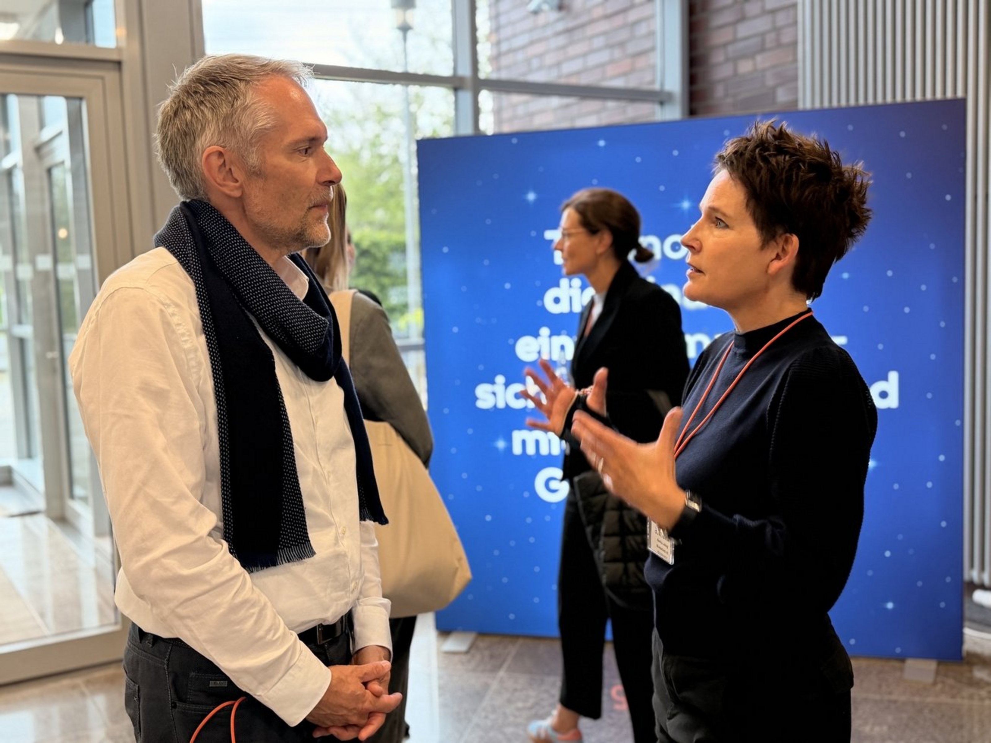 Zwei Personen stehen in einem Gebäude vor einer blauen Stellwand. Sie schauen sich an und sind im Gespräch. Im Hintergrund sind weitere Personen zu sehen.