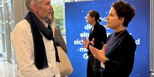 Zwei Personen stehen in einem Gebäude vor einer blauen Stellwand. Sie schauen sich an und sind im Gespräch. Im Hintergrund sind weitere Personen zu sehen.