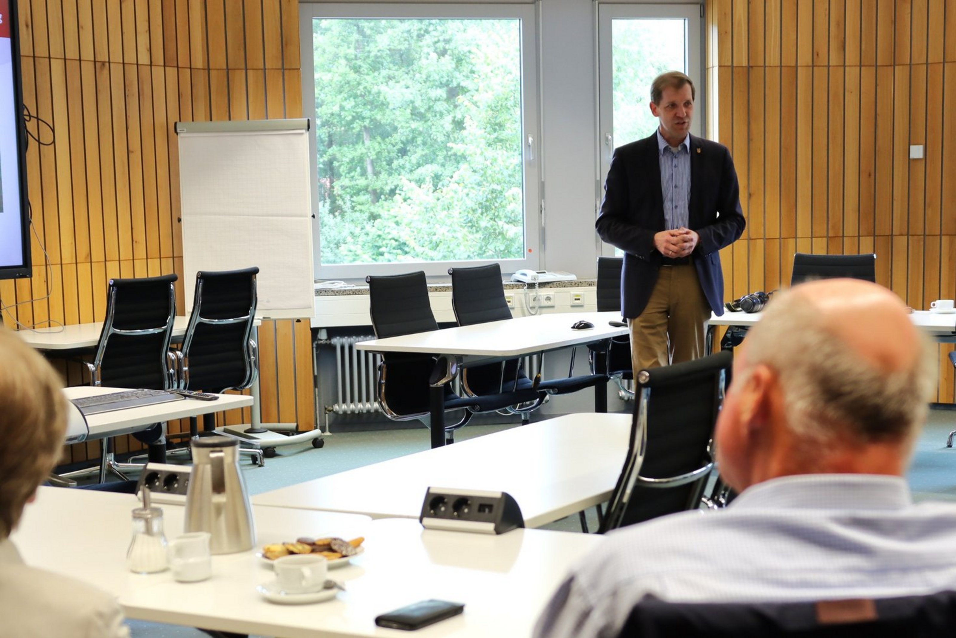 Landrat Dr. Christian Schulze Pellengahr informiert am 18. Juni über seine Aufgaben als Landrat und die Arbeit der Kreisverwaltung (Archivbild: Kreis Coesfeld) Ein Mann mit schwarzem Jackett, blauem Hemd und beiger Hose steht vor zwei sitzenden Personen in einem mit Holz vertäfeltem Raum. Die Hände hat er vor dem Bauch verschränkt. In dem Raum stehen Tischereihen mit schwarzen Stühlen. Vor den zwei sitzenden Personen, die von hinten zu sehen sind, stehen ein Keksteller, eine Thermoskanne, ein Salzstreuer und zwei Tassen.