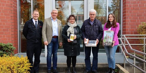 Auf einer Treppenstufe vor einem geklinkerten Gebäude stehen fünf Personen nebeneinander und schauen in die Kamera. Drei Personen halten Gegenstände in den Händen.
