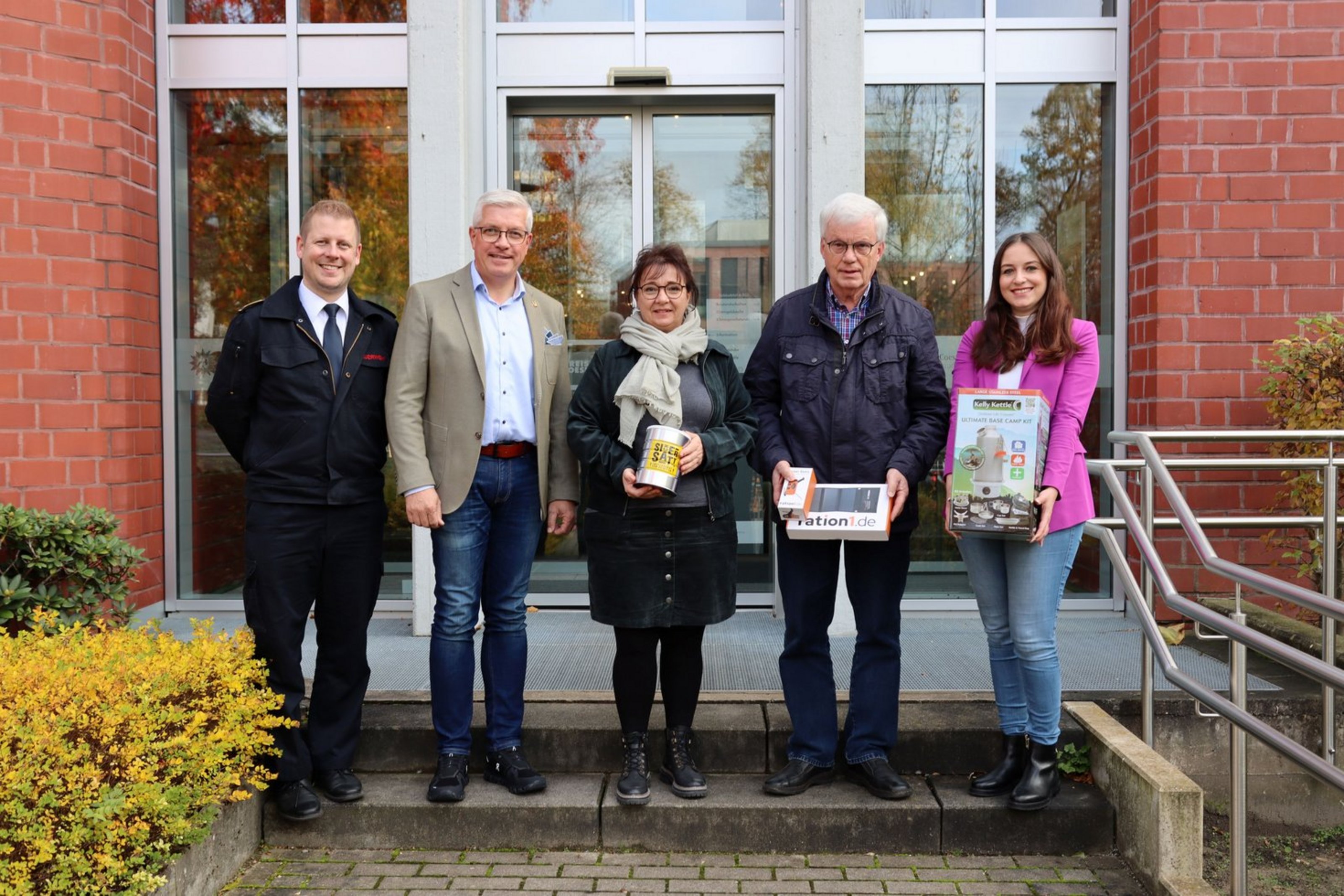 Auf einer Treppenstufe vor einem geklinkerten Gebäude stehen fünf Personen nebeneinander und schauen in die Kamera. Drei Personen halten Gegenstände in den Händen.