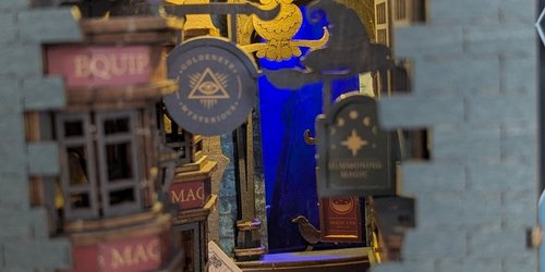 Durch einen Spalt in in einer Mauer sind ein Gässchen mit einem Laden, Schildern und Tieren zu sehen