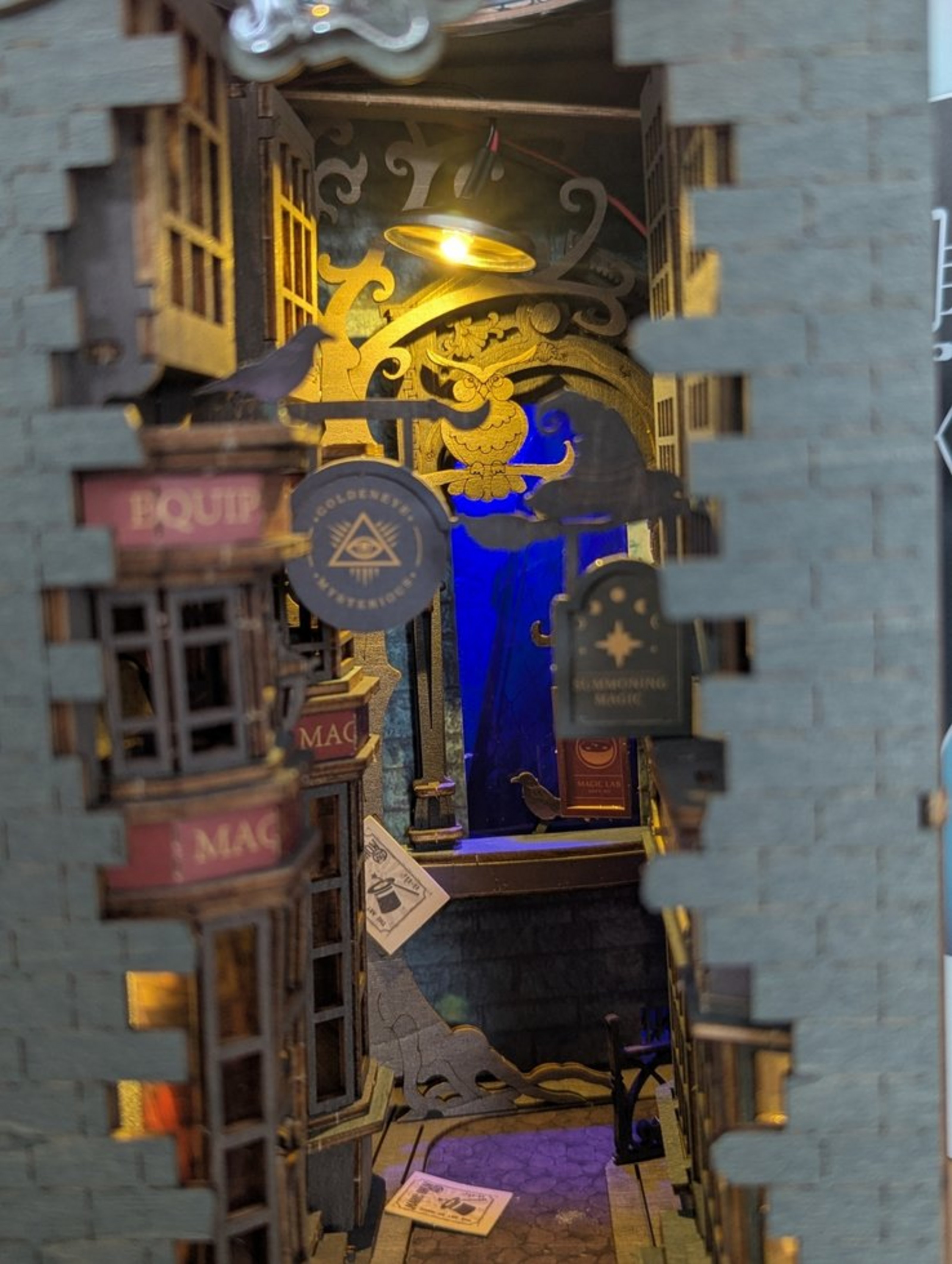 Durch einen Spalt in in einer Mauer sind ein Gässchen mit einem Laden, Schildern und Tieren zu sehen