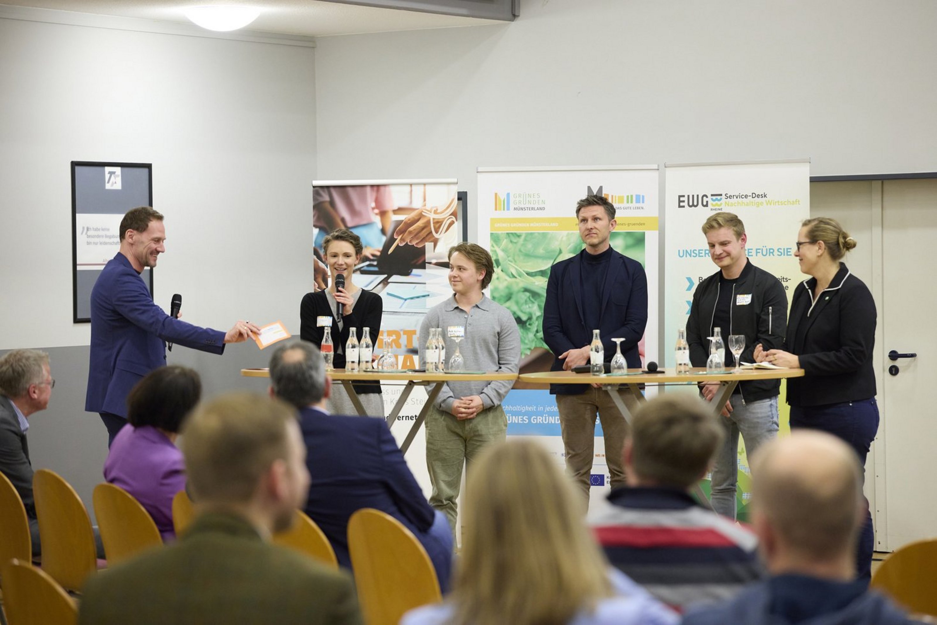 Sechs Personen stehen nebeneinander vor drei Aufstellern und vor einem sitzenden Publikum. Auf den Aufstellern steht "Grünes Gründen Münsterland". Zwei der Personen halten ein Mikrofon in der Hand.