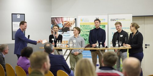 Sechs Personen stehen nebeneinander vor drei Aufstellern und vor einem sitzenden Publikum. Auf den Aufstellern steht "Grünes Gründen Münsterland". Zwei der Personen halten ein Mikrofon in der Hand.