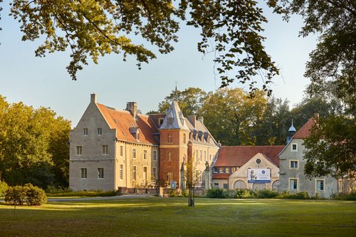 Hinter einer weitläufigen Rasenfläche ist ein dreiflügeliges Schloss mit rotem Ziegeldach inklusive kleinem Turm zu sehen. Umgeben ist das Schloss von Bäumen, der Himmel ist blau. 