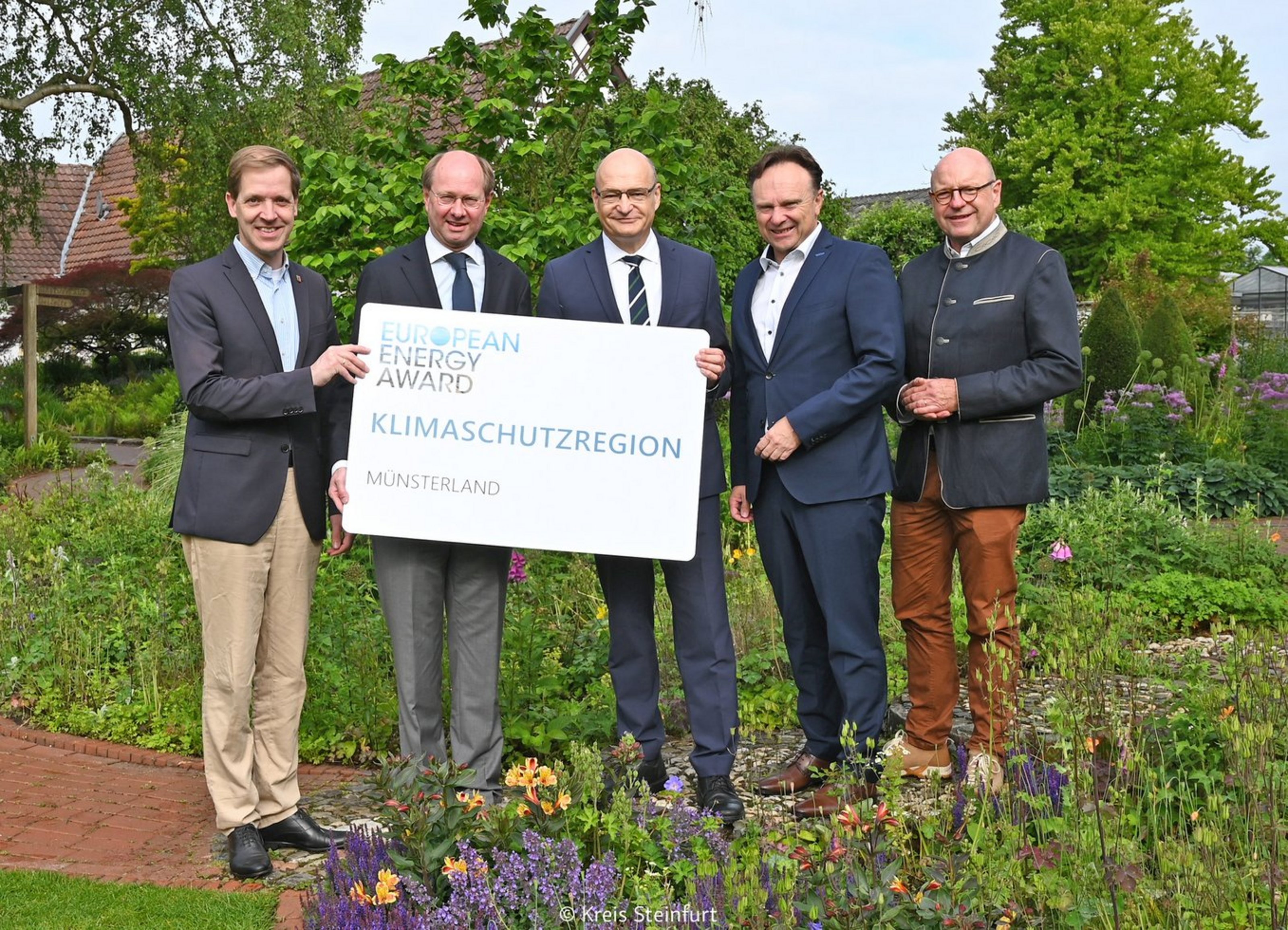 Das Münsterland ist Vorreiter-Region beim European Energy Award (Foto: Kreis Steinfurt). Fünf Männer stehen zwischen zwei Blumenbeten und halten ein Schild mit der Aufschrift "European Energy Award Klimaschutzregion Münsterland".