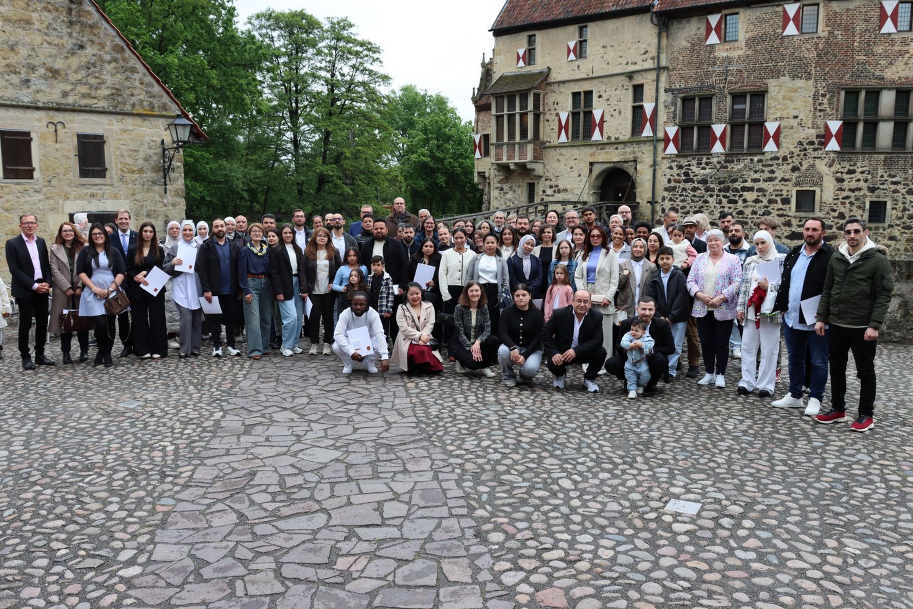 Eine Gruppe, bestehend aus 42 Personen steht auf einem gepflasterten Platz vor einer historischen Burg im Hintergrund.