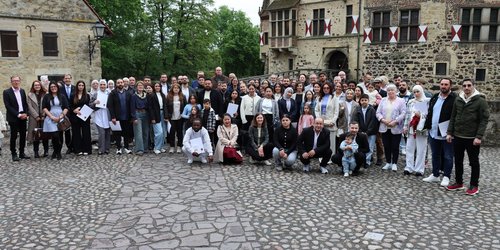 Eine Gruppe, bestehend aus 42 Personen steht auf einem gepflasterten Platz vor einer historischen Burg im Hintergrund.