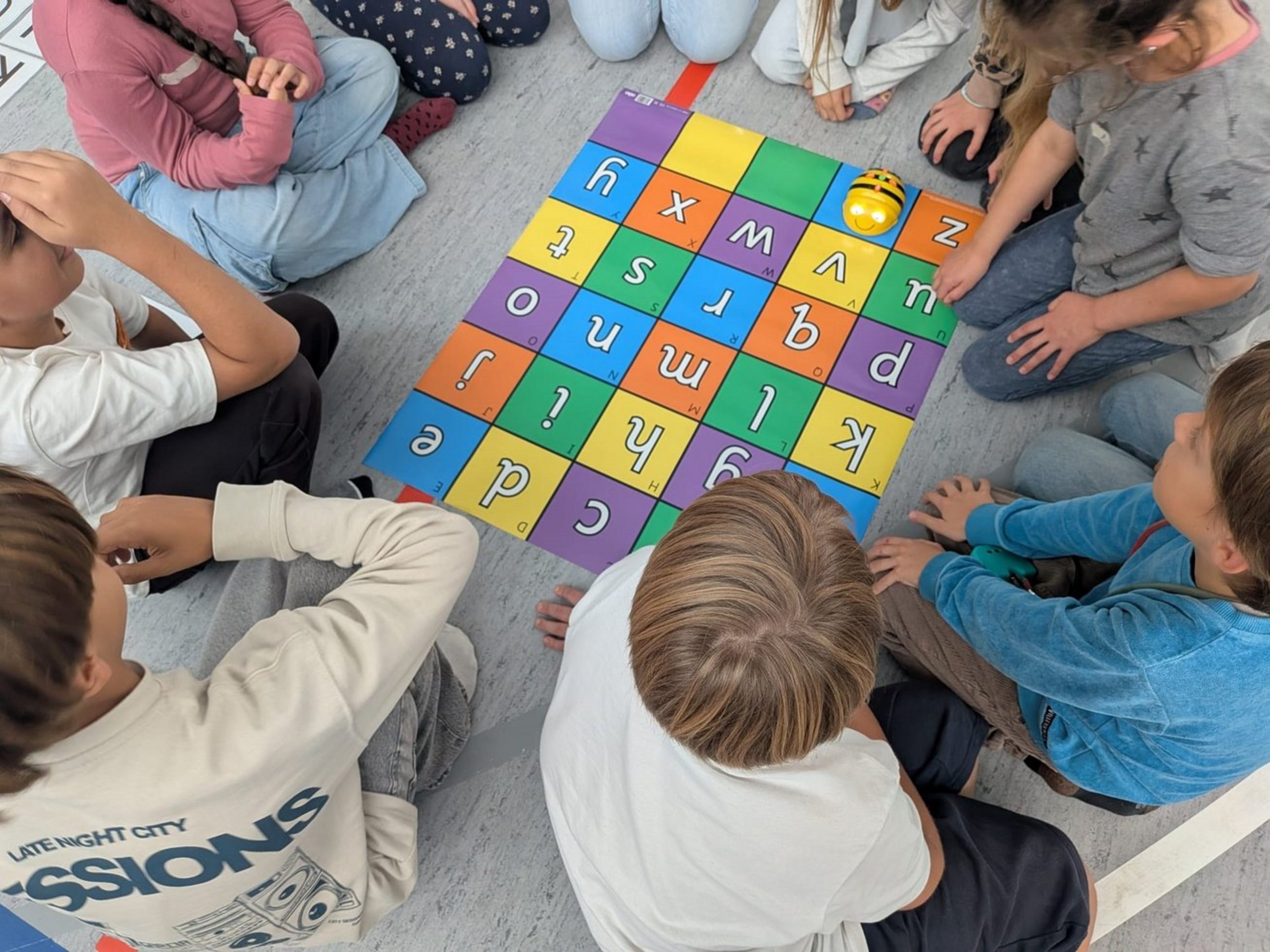 Kinder sitzen um eine bunte Buchstabenmatte und programmieren spielerisch einen Bee-Bot, der auf einem Buchstabenfeld steht.