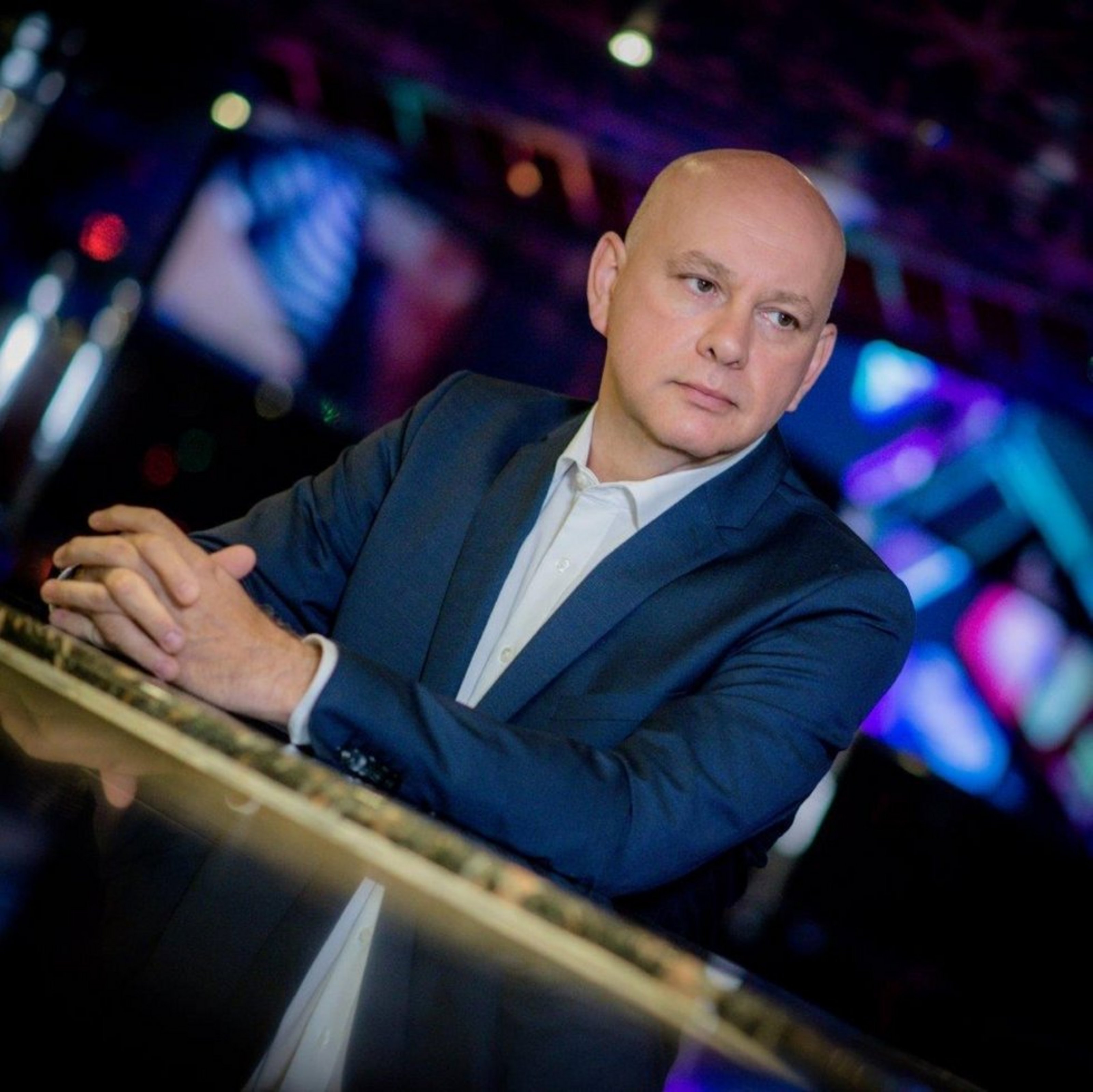 Ein Mann in blauem Jacket, hellem Hemd und Glatze stützt sich mit den Armen auf eine spiegelnde Platte. Im Hintergrund leuchtet es blau und lila. 