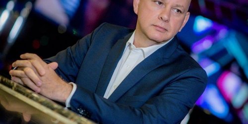 Ein Mann in blauem Jacket, hellem Hemd und Glatze stützt sich mit den Armen auf eine spiegelnde Platte. Im Hintergrund leuchtet es blau und lila. 