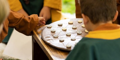 Ein Kind sitzt mit dem Rücken zur Kamera vor einem Spielbrett aus Holz im Freien auf Gras.