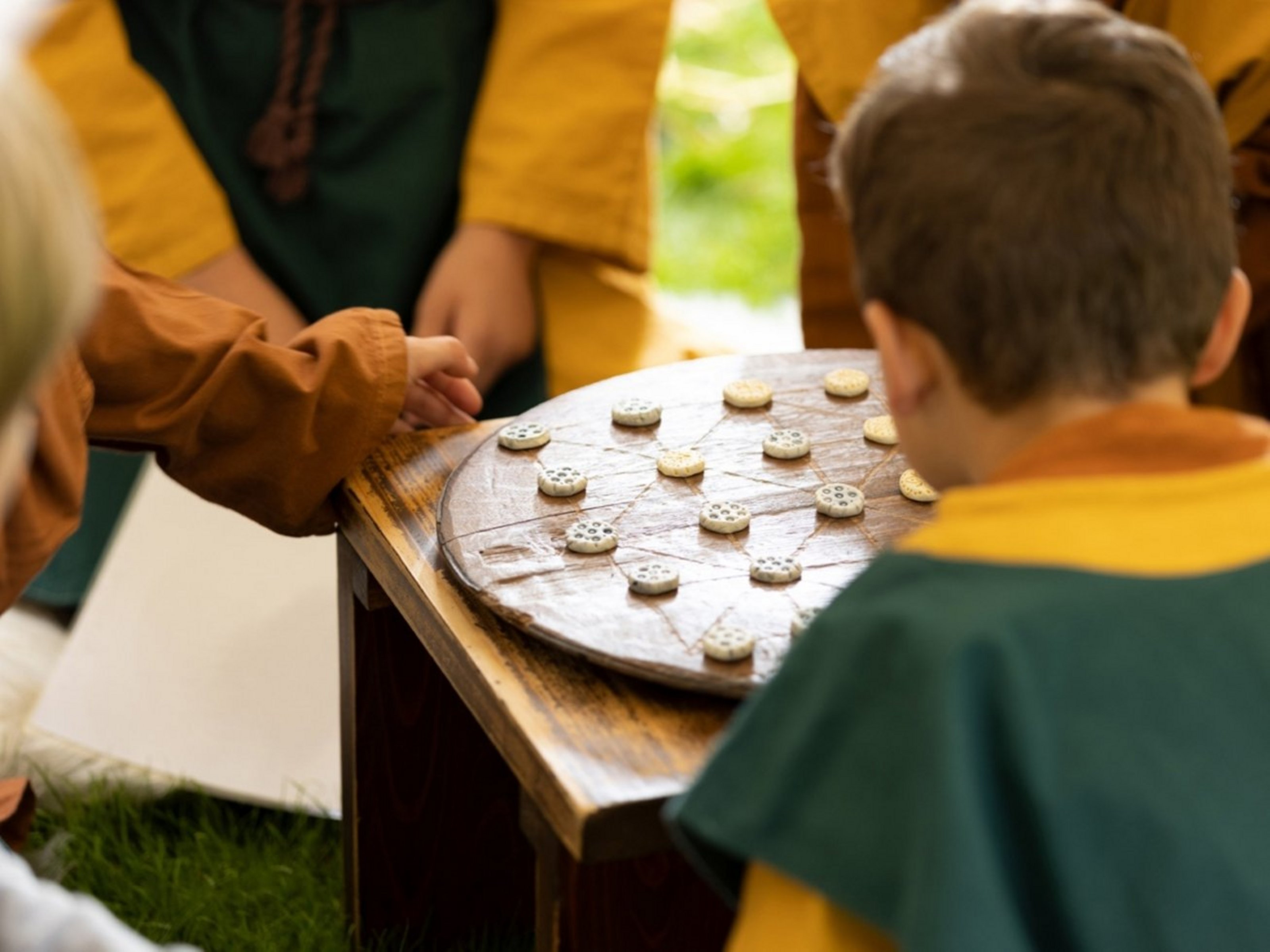 Ein Kind sitzt mit dem Rücken zur Kamera vor einem Spielbrett aus Holz im Freien auf Gras.