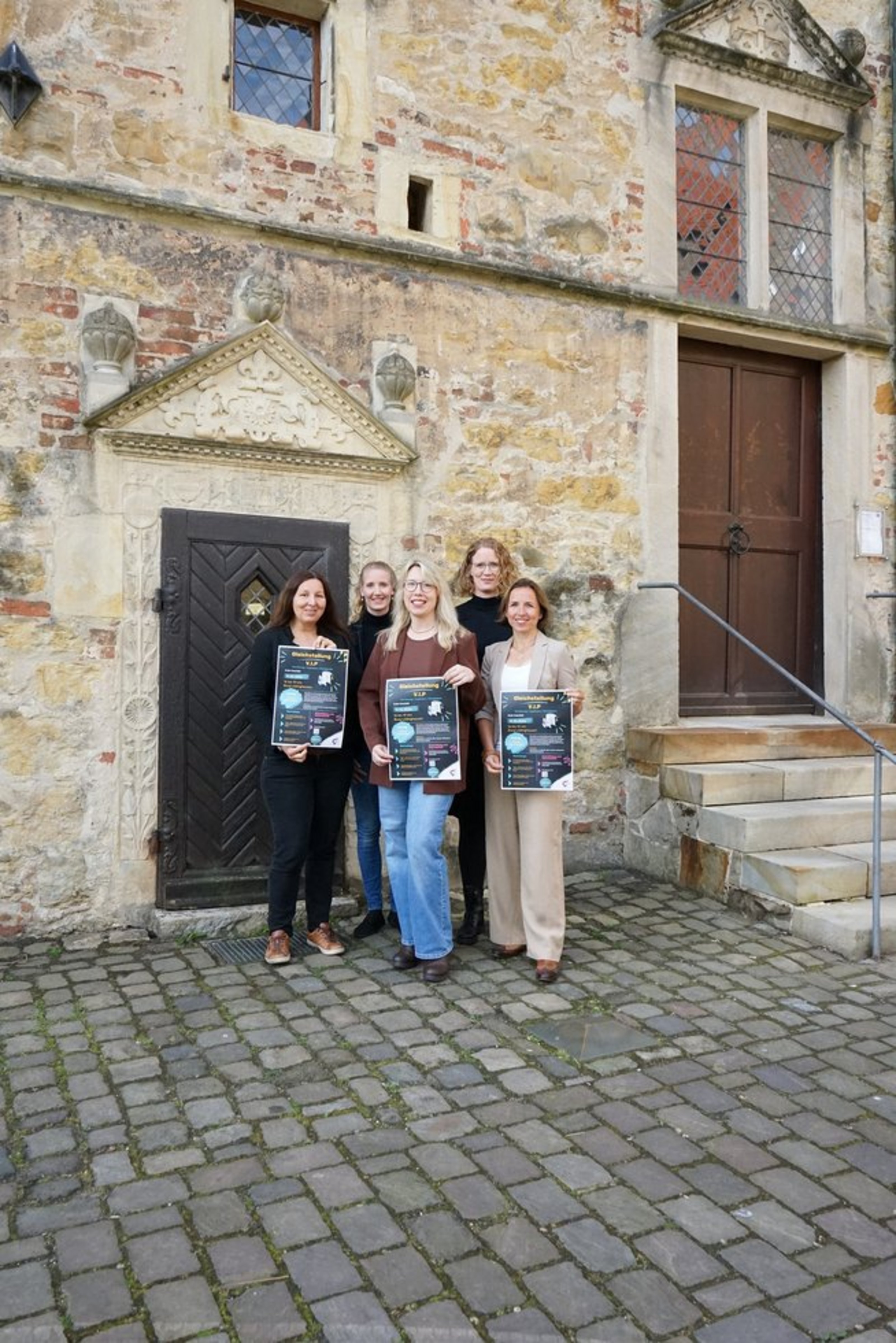 Fünf Frauen stehen in zwei Reihen nebeneinander, die drei vorderen halten ein Plakat jeweils in der Hand, auf dem "Gleichstellung V.I.P." sowie weiterer Text steht.Die Gruppe steht vor einem historischen Steingebäude mit Treppe und zwei Holztüren.