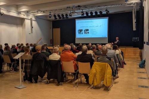 Vor einem Bildschirm sitzen viele Menschen auf Stühlen  im Publikum und blicken zu einem Mann, der mit Mikrofon vor ihnen steht. Der Fußboden ist aus Holz, die linke Wand weiß.