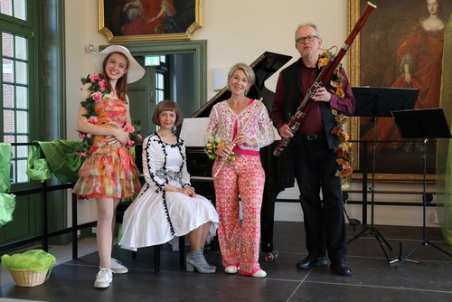 Vier Personen stehen in einem hellen Raum mit großen Fenstern und klassischer Einrichtung. Links steht ein junges Mädchen mit weißem Sonnenhut und einem bunten Kleid aus orangefarbenem, rosa und gelbem Stoff, geschmückt mit künstlichen Rosen. Daneben sitzt eine Frau mittleren Alters in einem eleganten weißen Kleid mit schwarzen Borten und silbernem Gürtel. Rechts davon steht eine weitere Frau in bunter Bluse aus Spitze und pink-gemusterten Schlaghosen, sie hält eine Flöte. Ganz rechts steht ein älterer Mann mit Brille, schwarzer Weste und einem Fagott, das mit künstlichen Herbstblättern dekoriert ist. Im Hintergrund steht ein Flügel mit Noten und Gemälden an den Wänden.