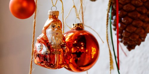Zwei rote Weihnachtskugeln, eine Weihnachtsmannfigur und ein Tannenzapfen hängen an einem Holzstück.