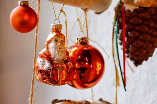 Zwei rote Weihnachtskugeln, eine Weihnachtsmannfigur und ein Tannenzapfen hängen an einem Holzstück.