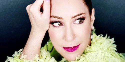 Eine Frau mit rötlicher Hochsteckfrisur und einem grünen, plüschigem Oberteil hat ihre rechte Hand an die Stirn gelegt und blickt nach rechts.