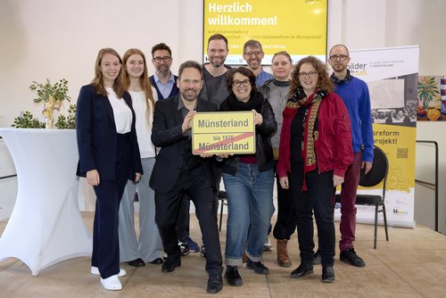 ine Gruppe von zehn Menschen steht gemeinsam vor einem gelben Plakat mit der Aufschrift „Herzlich willkommen! Auftaktveranstaltung ‚Schilderwechsel – 50 Jahre Gebietsreform im Münsterland‘“. Zwei Personen in der Mitte halten ein gelbes Ortsschild mit dem Text „Münsterland bis 1975 / Münsterland“ in den Händen. Alle Personen schauen freundlich in die Kamera. Im Hintergrund sind Dekorationen, weitere Plakate und ein Stehtisch mit Blumenschmuck zu sehen. Die Veranstaltung findet in einem hellen Innenraum statt.