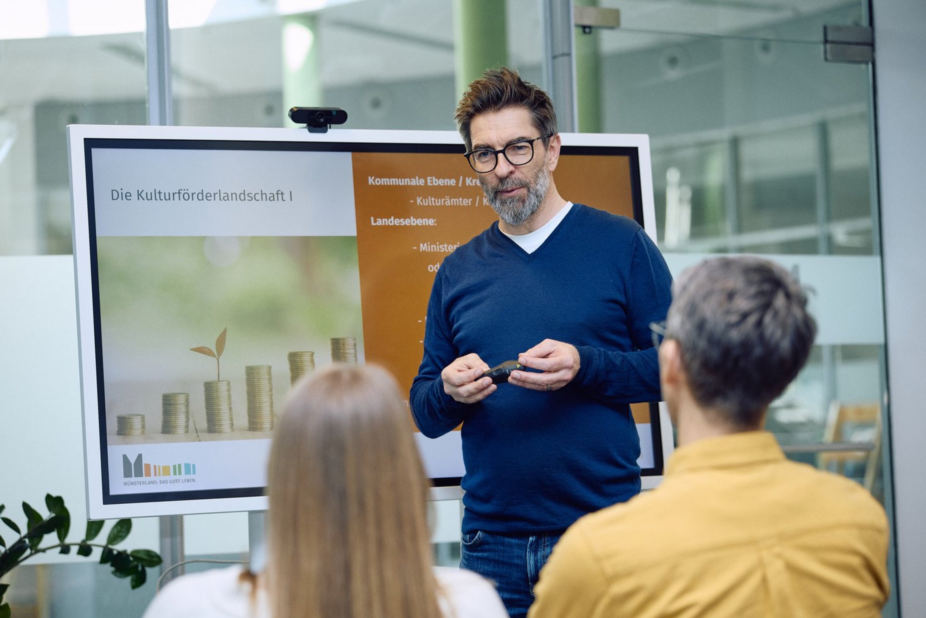 Referent erklärt vor Teilnehmenden die Grundlagen der Kulturförderung in Nordrhein-Westfalen anhand einer Präsentation auf einem Bildschirm.