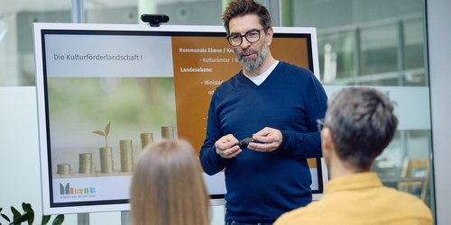 Referent erklärt vor Teilnehmenden die Grundlagen der Kulturförderung in Nordrhein-Westfalen anhand einer Präsentation auf einem Bildschirm.
