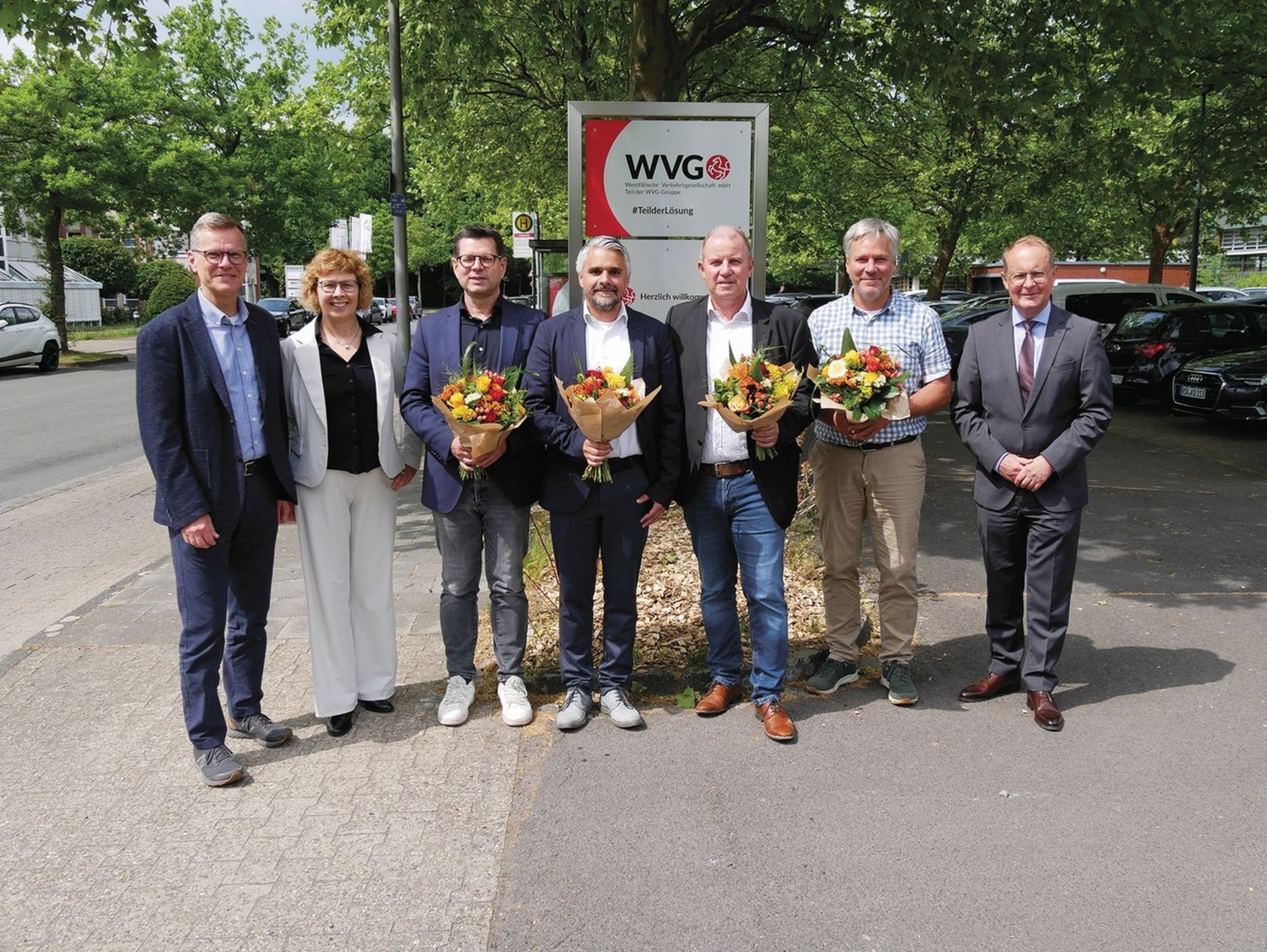 Sieben Personen stehen vor einem WVG-Schild; vier Männer in der Mitte halten Blumensträuße.