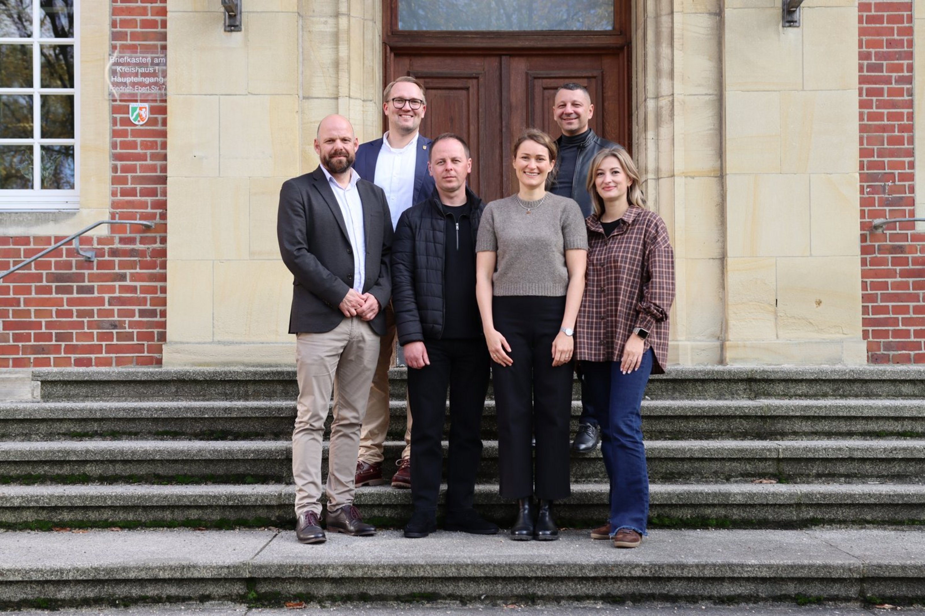 Eine Gruppe, bestehend aus sechs Personen, steht auf der Treppe vor der EIngangstür eines Backsteingebäudes und blickt in die Kamera.
