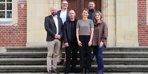 Eine Gruppe, bestehend aus sechs Personen, steht auf der Treppe vor der EIngangstür eines Backsteingebäudes und blickt in die Kamera.