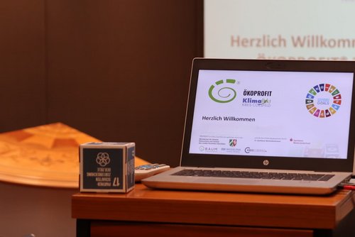 Laptop mit ÖKOPROFIT®-Präsentation auf einem Tisch; im Hintergrund ist „Herzlich Willkommen“ zu sehen – Symbolbild für eine Informationsveranstaltung.