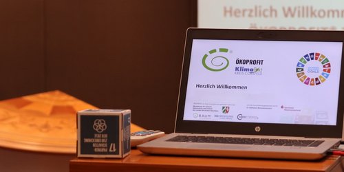 Laptop mit ÖKOPROFIT®-Präsentation auf einem Tisch; im Hintergrund ist „Herzlich Willkommen“ zu sehen – Symbolbild für eine Informationsveranstaltung.