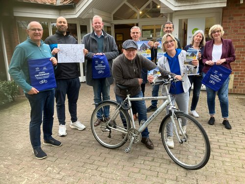 Eine Gruppe von neun Personen steht vor einem Gebäude mit einem überdachten Eingang. Alle lächeln in die Kamera und halten verschiedene Materialien rund ums Radfahren in der Region – darunter Flyer, Broschüren, Karten und blaue Stoffbeutel mit der Aufschrift „COOL UNTERWEGS“. Im Vordergrund steht ein silberfarbenes Fahrrad der Marke Gudereit. Ein Mann lehnt sich lässig an das Rad. Das Wetter wirkt mild, alle Personen tragen Freizeitkleidung oder legere Jacken. Die Szene vermittelt eine freundliche, aktive Stimmung rund um das Thema Fahrradtourismus.