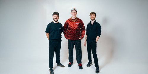 Drei Männer stehen nebeneinander vor weißem Hintergrund und haben die Händen in ihren Hosentaschen.  Zwei tragen blaue, einer trägt ein rotes Oberteil.