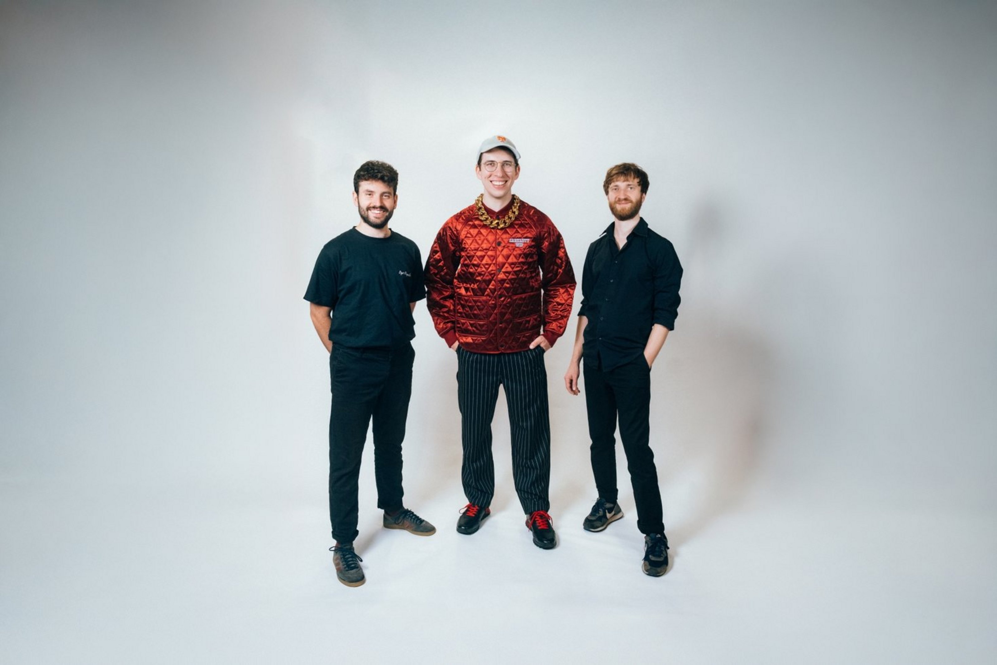 Drei Männer stehen nebeneinander vor weißem Hintergrund und haben die Händen in ihren Hosentaschen.  Zwei tragen blaue, einer trägt ein rotes Oberteil.