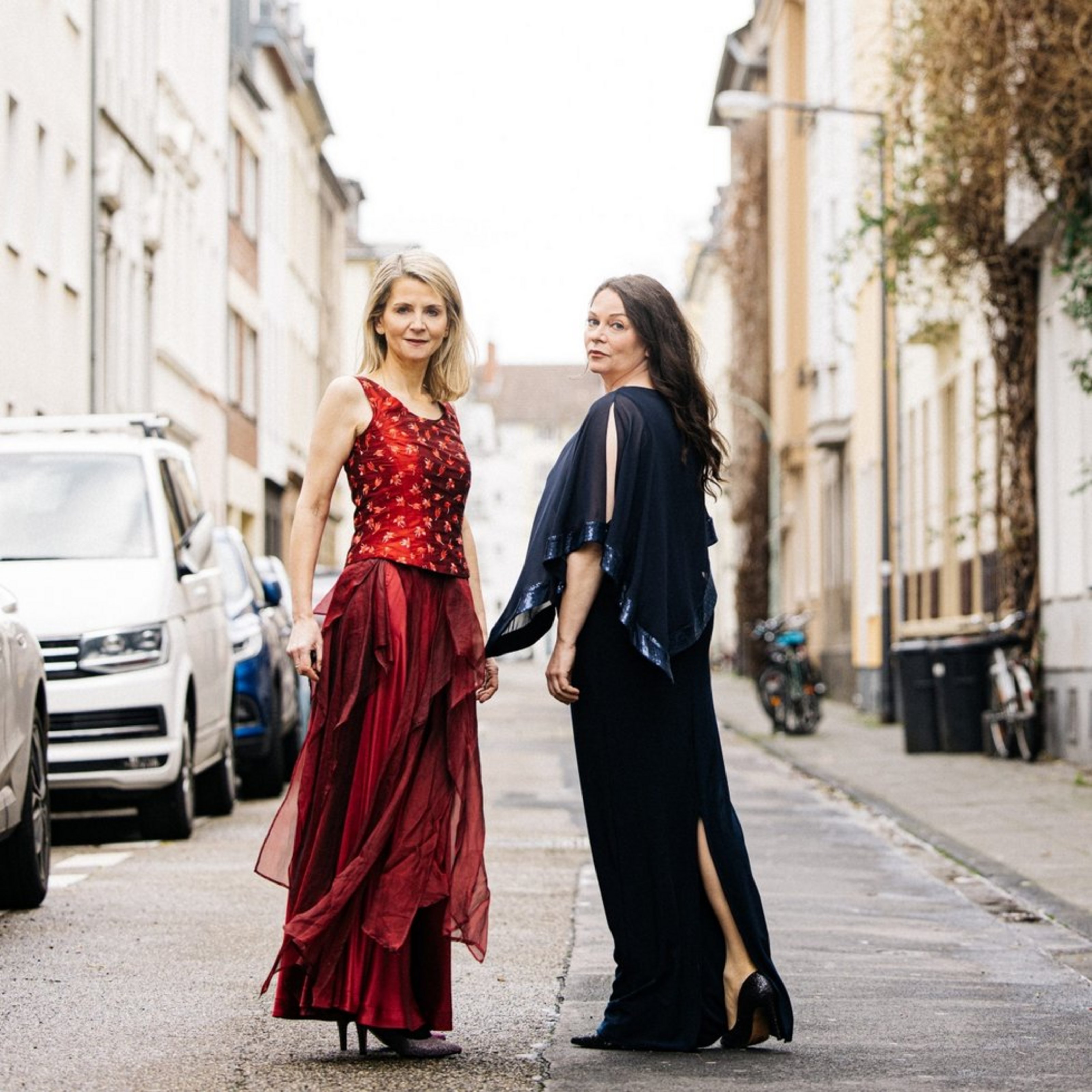 Zwei Frauen stehen auf einer städtischen Straße. Eine trägt ein rotes Kleid und schaur in die Kamera, die andere trägt blaue Kleidung, iläuft von der kamera weg und schaut dabei über die Schulter zurück. Am Straßenrand parken Autos.
